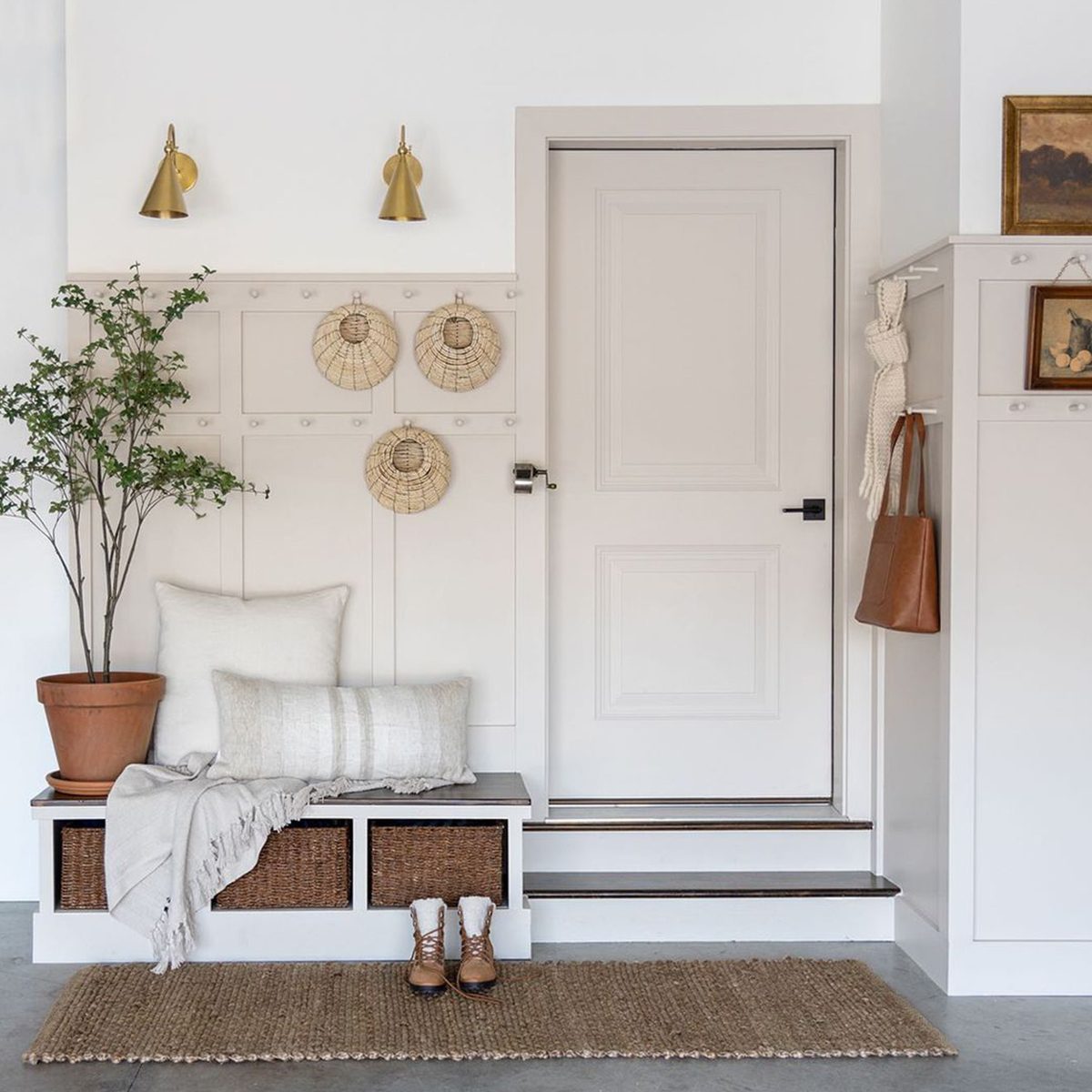 A bench with pillows sits beside a door, featuring decorative wall hangings and indoor plants in a minimalist, well-lit entryway.