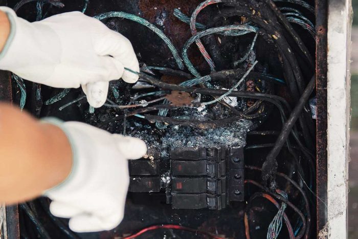 A person in gloves inspects a damaged electrical panel, pointing at burnt wires amidst a tangled mass of blackened cables and debris.