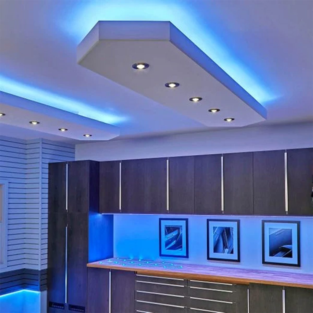 A modern kitchen features sleek cabinets and illuminated ceiling lights. Blue ambient lighting enhances the atmosphere, while framed art pieces decorate the walls.