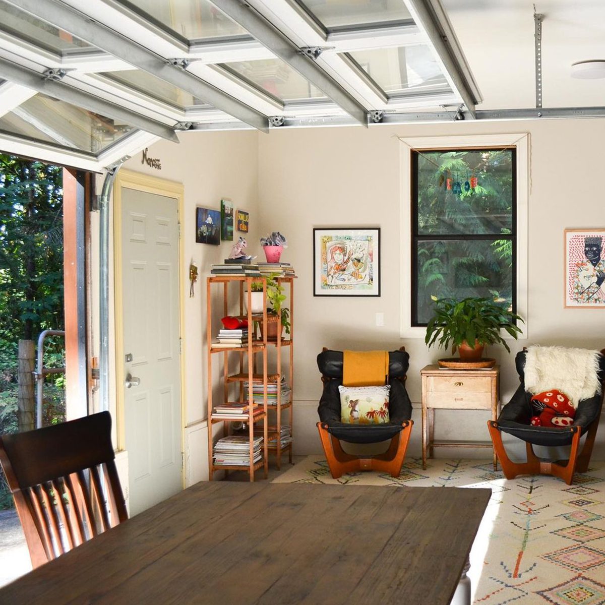 A dining table sits in a bright garage, featuring two cozy chairs, a plant, bookshelves, and wall art, with an open garage door revealing greenery outside.