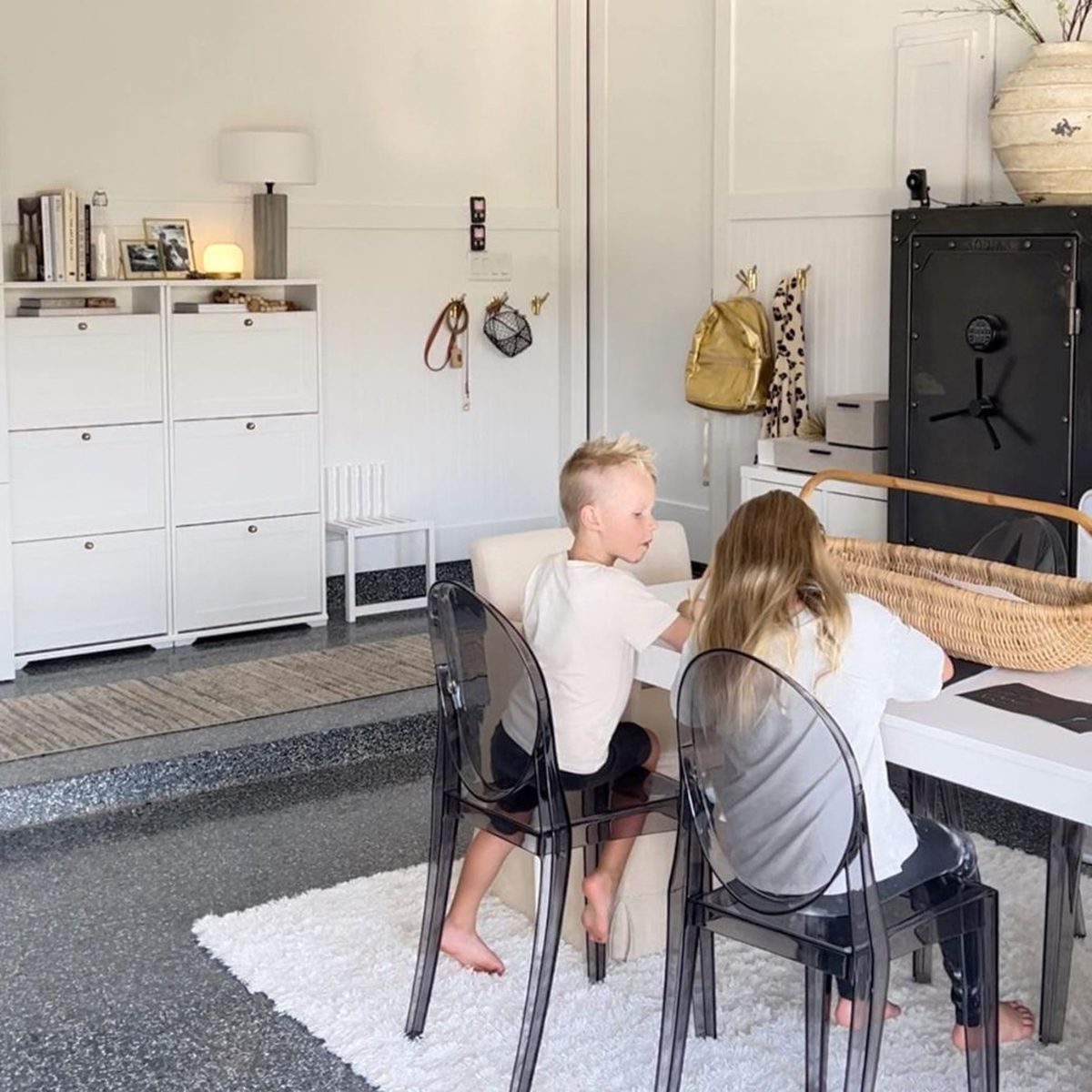 Two children sit at a table, focused on activities. A modern room features white furniture, transparent chairs, and decorative items in a bright, minimalist setting.