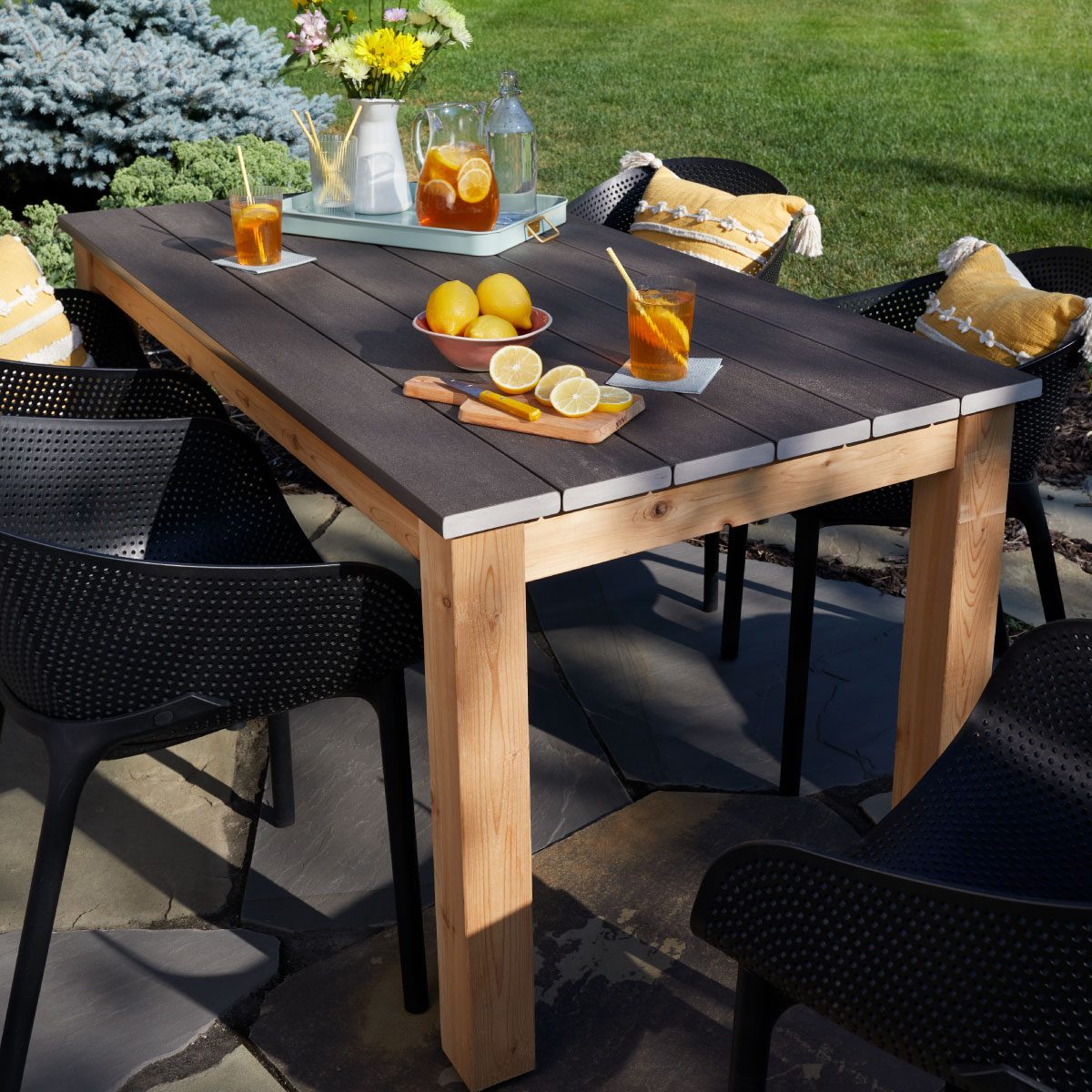 A wooden table showcases drinks and lemons, surrounded by black chairs, on a sunny outdoor patio with green grass and a blue tree backdrop.
