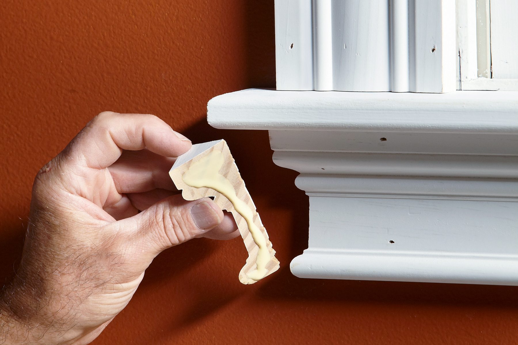 A person holding a small piece of wood with a decorative design alongside a white window frame against a rust-colored wall, demonstrating where the piece will be attached.