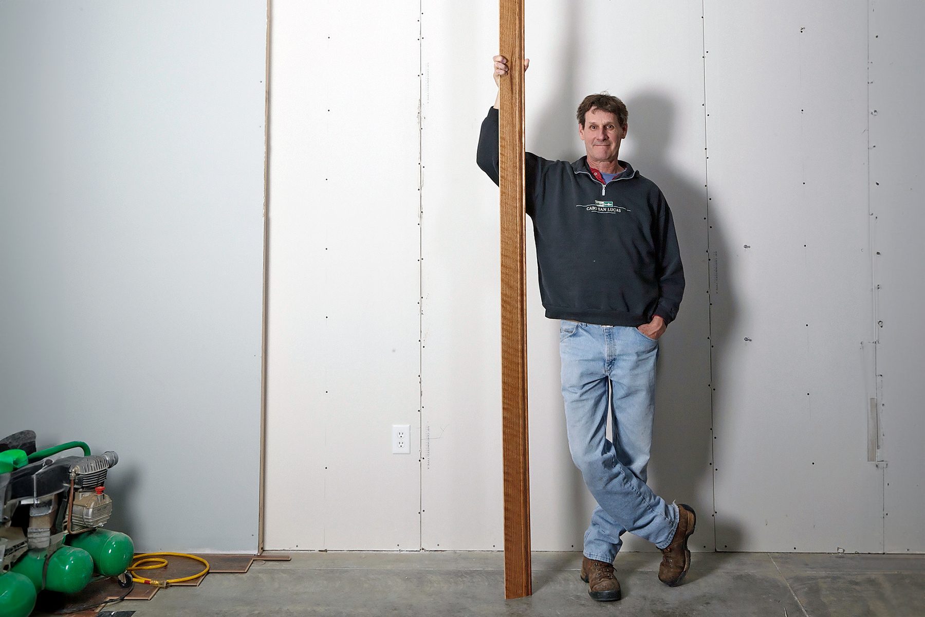 A man in a dark hoodie and jeans stands in a room with unfinished walls, holding a long wooden plank vertically. There are various tools and green objects on the ground to his left.