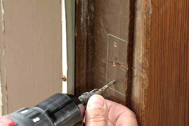 A person using a power drill to create a hole in a wooden door frame. The hand is steady, guiding the drill bit into the wood, showing a close-up of the DIY task.