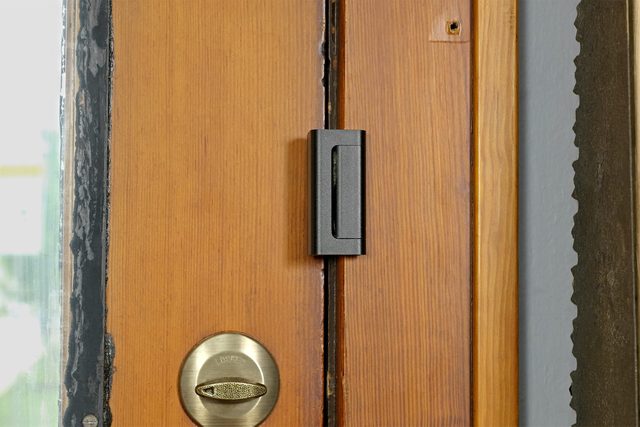A close-up of a wooden door features a brass deadbolt lock on the left side of the image and a black door sensor mounted vertically in the center. The edge of the door shows wear, revealing layers underneath the paint.