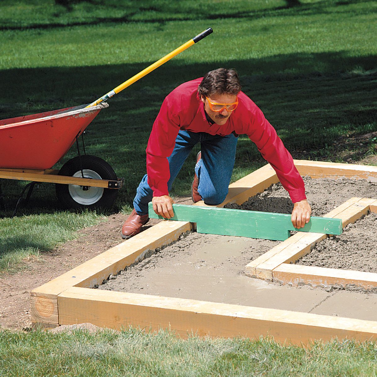 A person wearing a red shirt and blue jeans is leveling concrete within a wooden frame using a green tool. A red wheelbarrow is nearby on a grassy area. The background features a green lawn and partial shade.