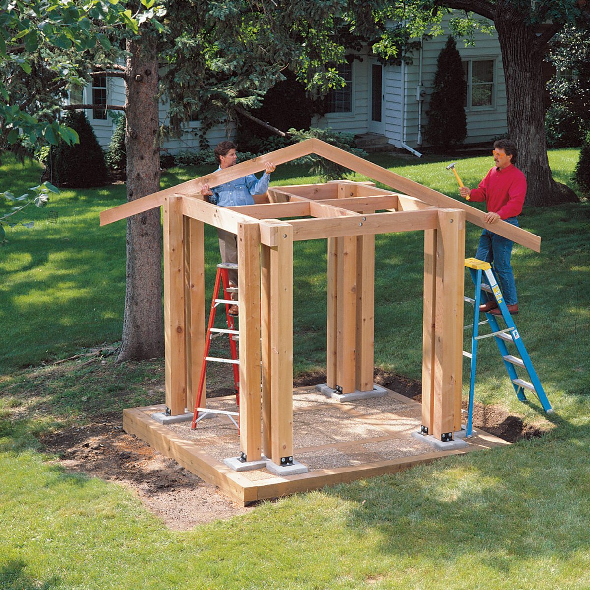 Two people are constructing a wooden shed frame outdoors. The frame consists of vertical and horizontal beams forming the structure's skeleton. One person on a red ladder and another on a blue ladder work on attaching pieces.