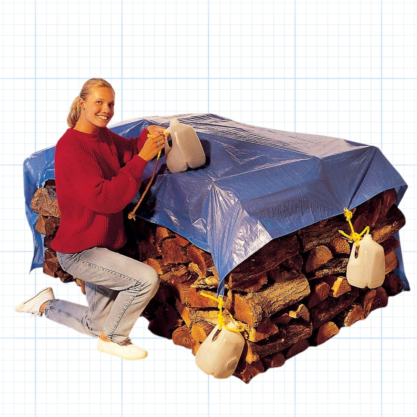 A person with light hair, wearing a red sweater and light blue jeans, is kneeling next to a stack of firewood. The firewood is covered with a blue tarp that is weighed down by white jugs tied to the tarp