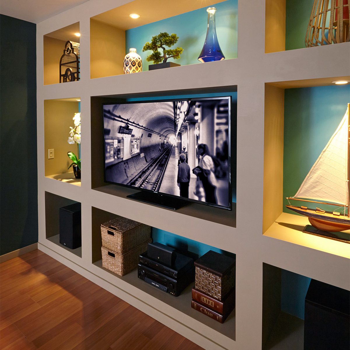 A modern living room setup featuring a built-in wall unit with multiple compartments displaying decorative items, such as a ship model, a vase, and baskets. A flat-screen TV is mounted in the center, showing a black-and-white image of a tunnel.