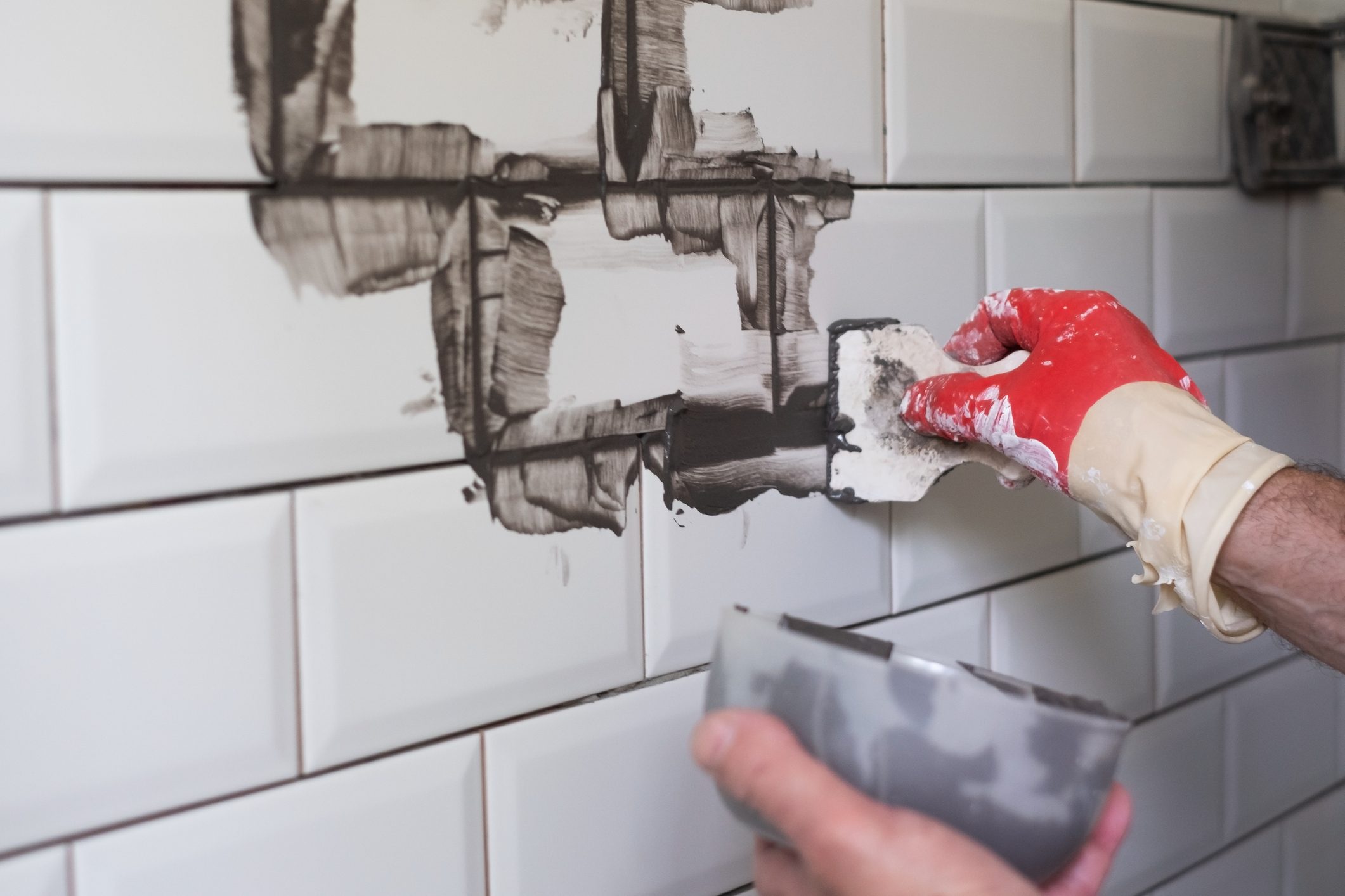 Worker applies grey grout at white tiles with rubber trowel.
