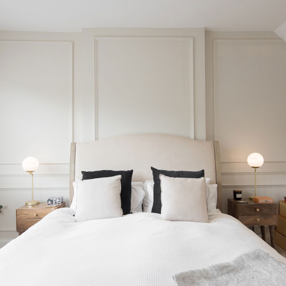 A general interior view of a elegant bedroom painted in natural cream colours, with wall paneling, bed, cushions, wooden night table with drawers, gold, brass table lamps with white glass globe shades, blanket, beige carpet, storage boxes, window with venetian blinds, vase within a home.