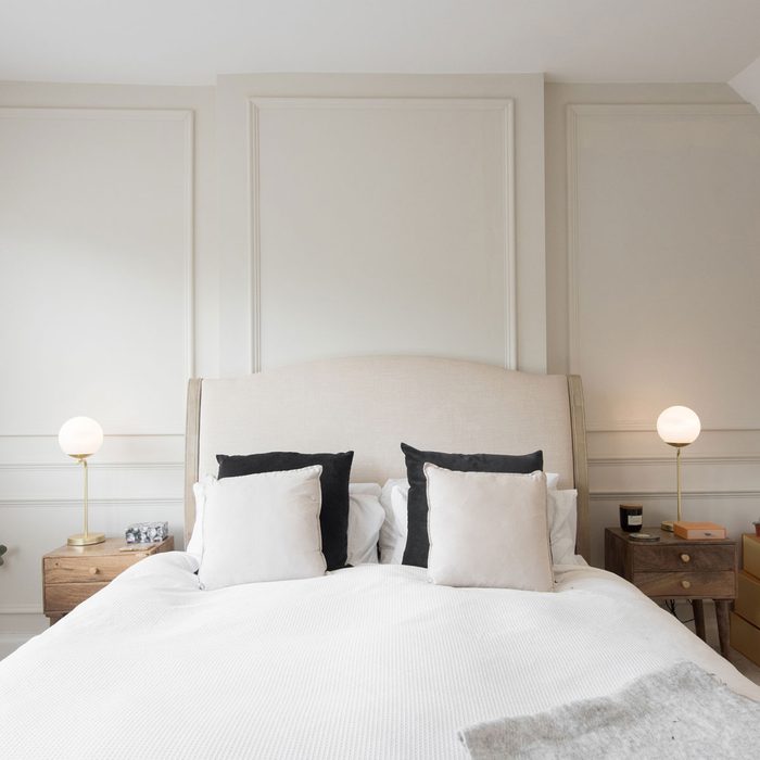 A general interior view of a elegant bedroom painted in natural cream colours, with wall paneling, bed, cushions, wooden night table with drawers, gold, brass table lamps with white glass globe shades, blanket, beige carpet, storage boxes, window with venetian blinds, vase within a home.