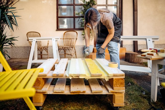 A person wearing an apron paints wooden boards on a makeshift table outdoors. The area is set with chairs, plants, and a window in the background, suggesting a garden or patio.