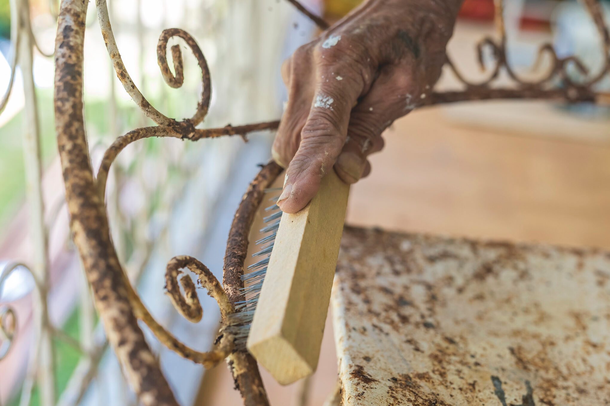 A man uses a steel brush to scrape off old paint and rust from an old wrought iron chair. Furniture repair and restoration concept.