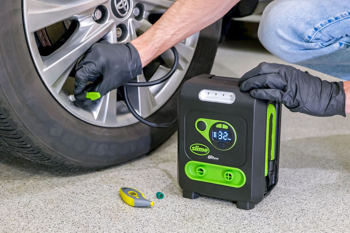 A person is using an air compressor to inflate a car tire; the person is wearing black gloves; a tire pressure gauge is also visible on the ground