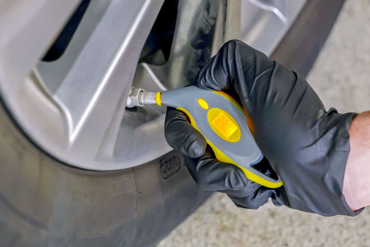 a person checking the tire pressure of a car; they are wearing black gloves and using a digital tire pressure gauge; the tire is visible in the background