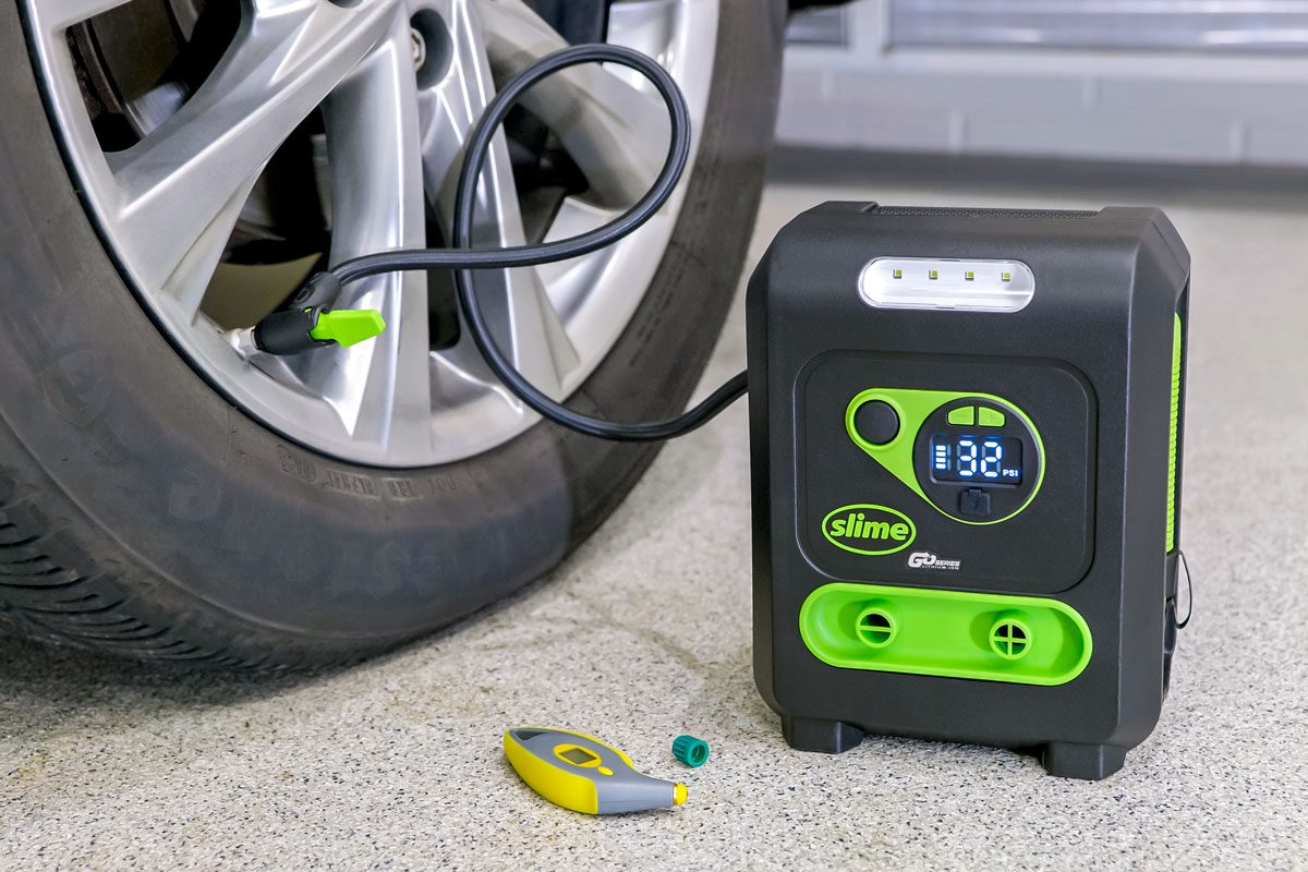 a car tire with a tire pressure gauge attached to its valve stem; the gauge is connected to a portable tire inflator, which is sitting on the ground near the tire;