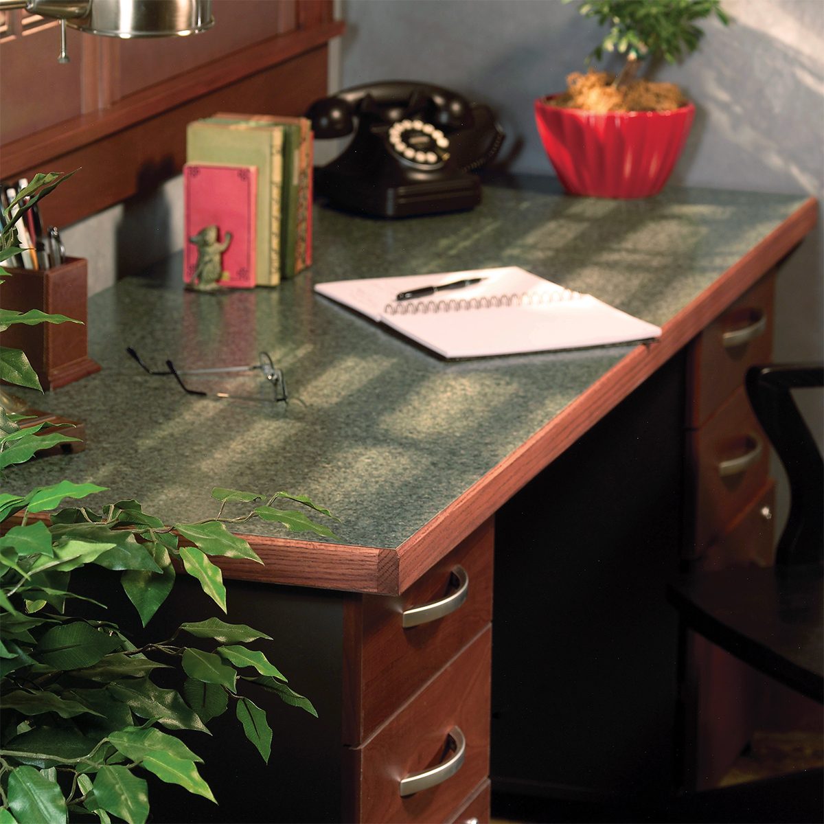 A home office desk with a green surface. Items include a retro black rotary phone, green and red books, a notepad, and eyeglasses. A red potted plant is in the corner. Sunlight casts shadows across the desk and a chair is partially visible.