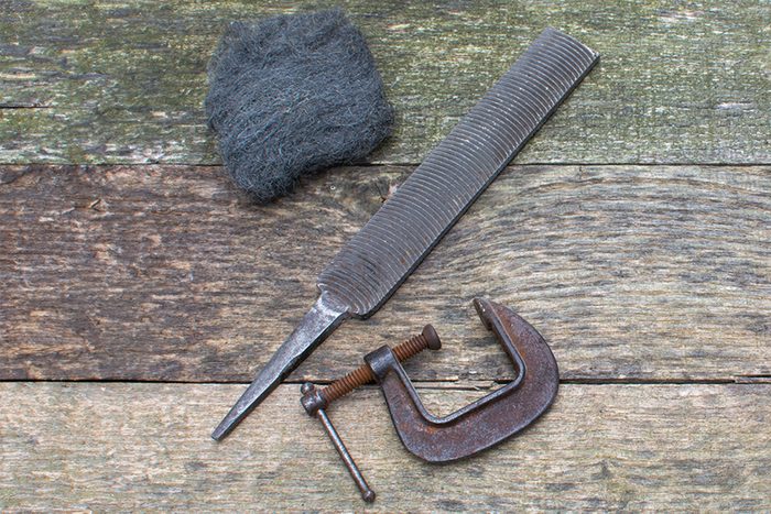 A piece of steel wool, a metal file, and a rusty C-clamp are laid out on a weathered wooden surface. The tools show signs of age and wear.