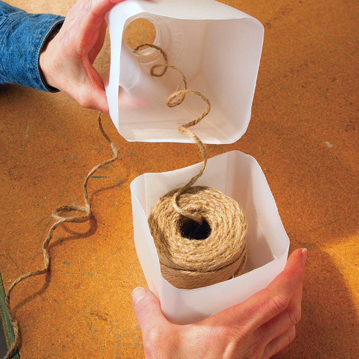 Hands holding a homemade twine dispenser made from a repurposed plastic container. The container is cut open with twine inside, and a piece of twine is threaded through a hole for easy access. The background is a wooden surface.