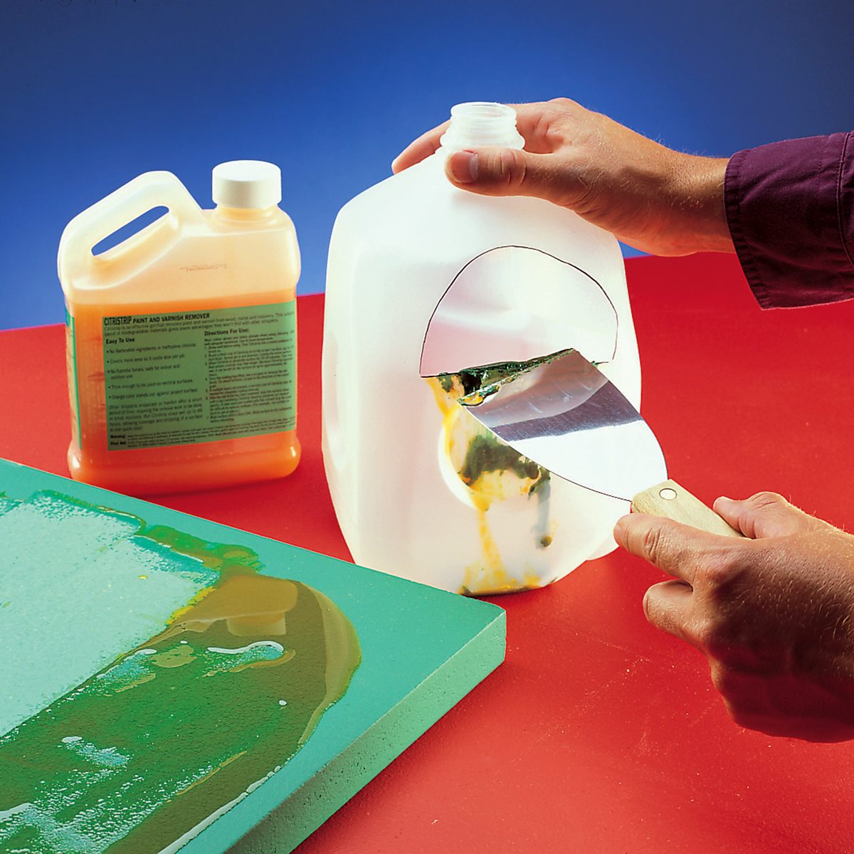 Two hands cut a large white plastic jug in half with a hand saw. Another orange liquid container with a label is in the background. The scene is on a red surface, with green material partially painted with adhesive in front.