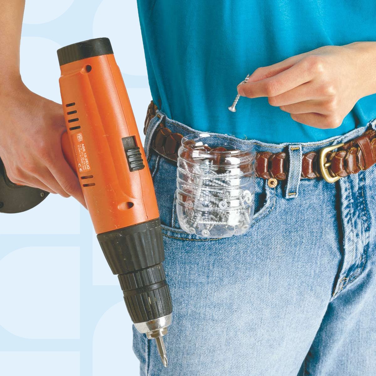 A person in a blue shirt and jeans holds a nail in one hand and an orange drill in the other. A clear plastic bottle, used as a tool holder, is attached to their belt, containing nails and screws.