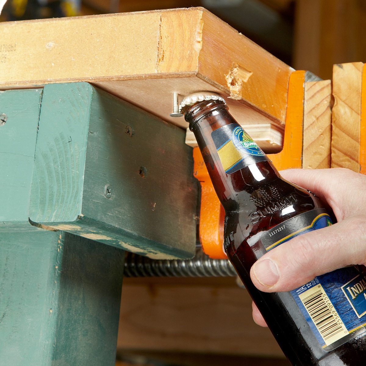 A hand using a workbench as a bottle opener to open a brown glass beer bottle. The top of the bottle is being pushed against the edge of a wooden bench clamped in place. The scene is in a workshop environment.
