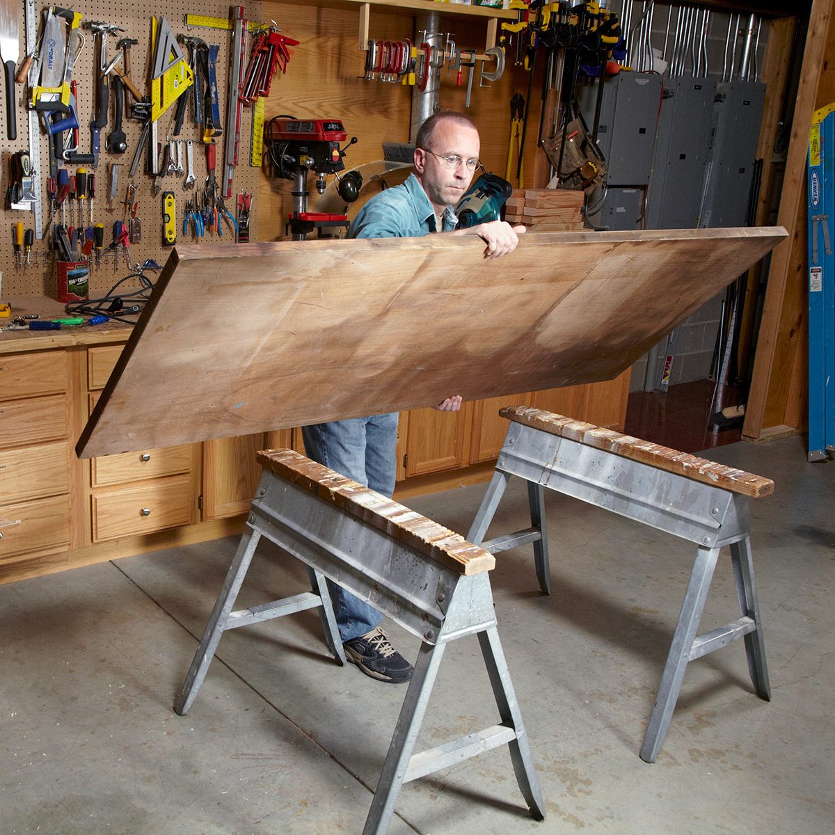 A person in a workshop setting is lifting a large wooden board onto two sawhorses. The background shows various tools hanging on the wall and a wooden workbench. They are wearing casual clothing and glasses.