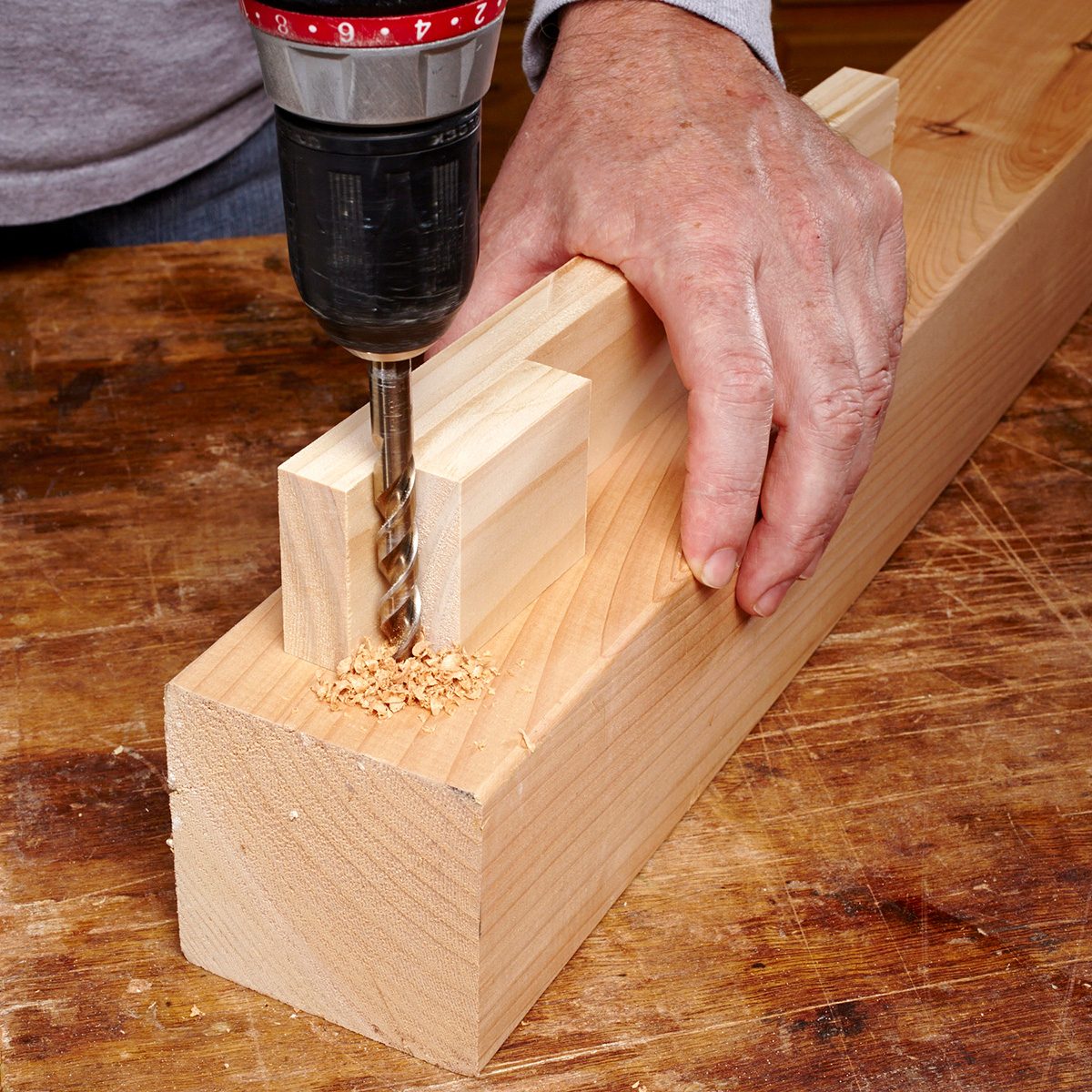 A person using a drill to create a hole in a piece of wood, clamped onto a wooden workbench. Sawdust is visible around the drill bit as it penetrates the rectangular wooden block. The person