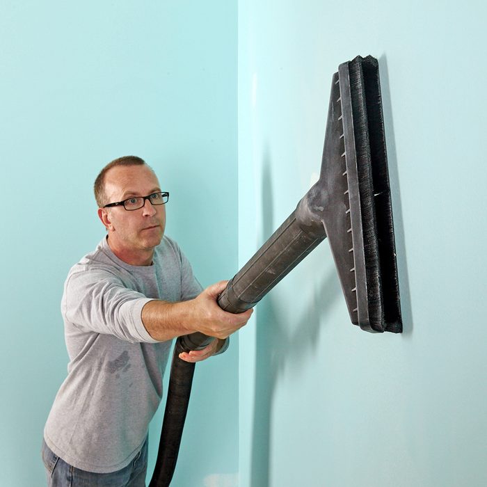A person with glasses uses a large vacuum attachment to clean a light blue wall. They are wearing a gray long-sleeve shirt and focusing on their task in a room with matching walls.