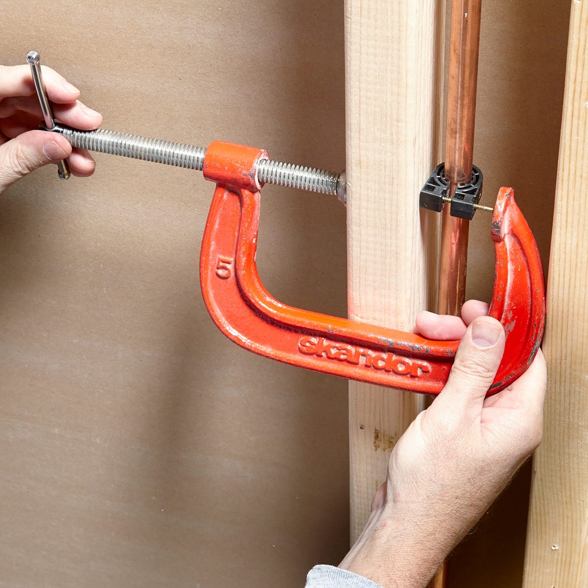 Hands using an orange C-clamp to hold copper piping against a wooden beam. The word