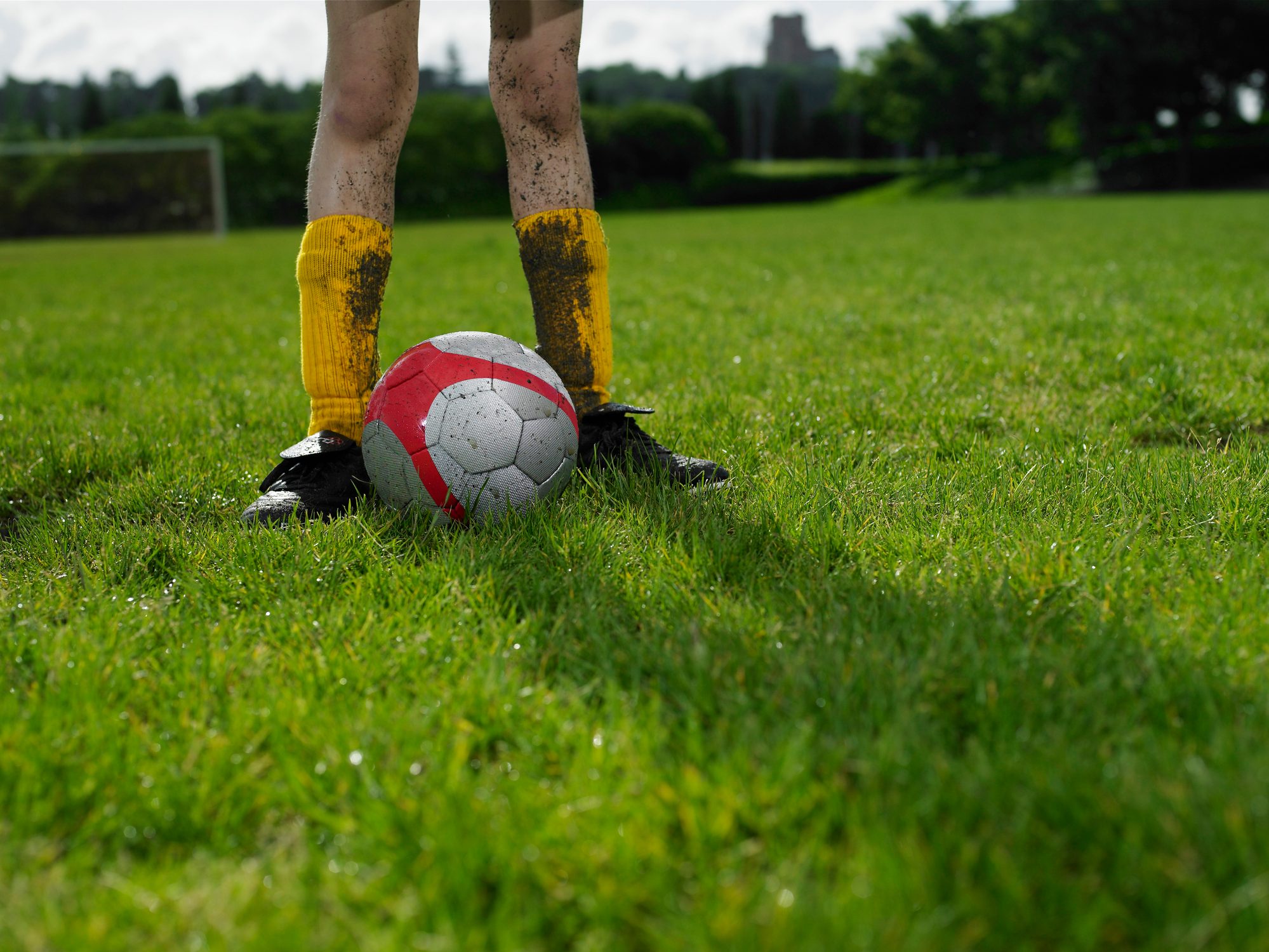 Person with dirty yellow shin guards stands behind a soccer ball on a grassy field