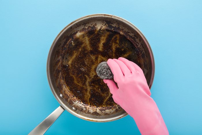 Woman hand in rubber protective glove holding steel wool and washing stainless frying pan with burn grease stain on blue table background. Closeup. Pastel color. Point of view shot. Top down view.
