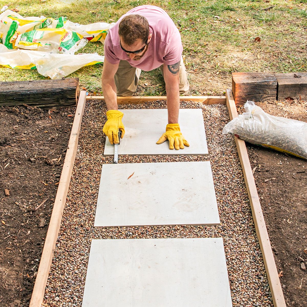 a person in a pink shirt and sunglasses carefully installing or aligning white cement boards or pavers in a linear pattern over a gravel-filled wooden frame; they