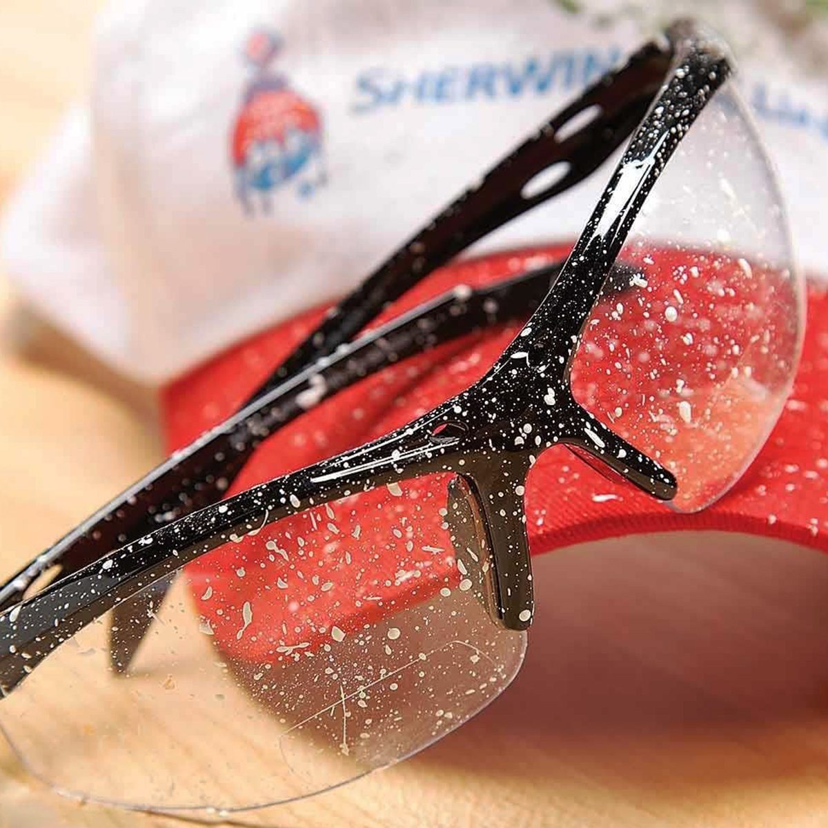 Safety glasses speckled with white paint resting on a red and white cap. The focus is on the glasses, showing paint splatters on the lenses and frames. The cap is partially visible in the background.