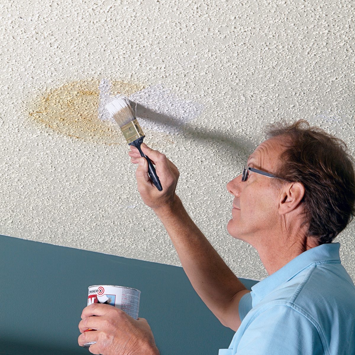 A person wearing glasses paints over a stain on a textured white ceiling with a brush, holding a small can of paint. The wall below is painted teal. The stain appears to be a water mark.