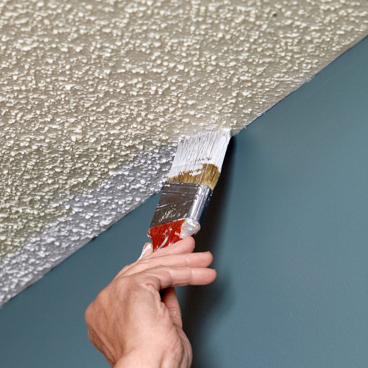 A hand is using a paintbrush to carefully apply paint along the edge where a textured ceiling meets a smooth, teal-colored wall. The ceiling is painted white, while the wall is a deep teal shade.