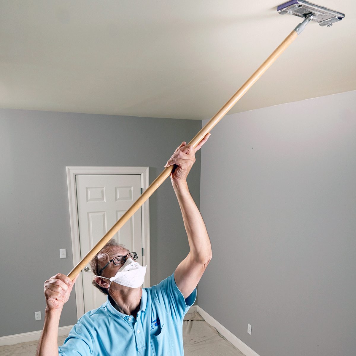 A person wearing a mask and safety glasses uses a long-handled tool to clean or paint a ceiling in a room with gray walls and a white door.