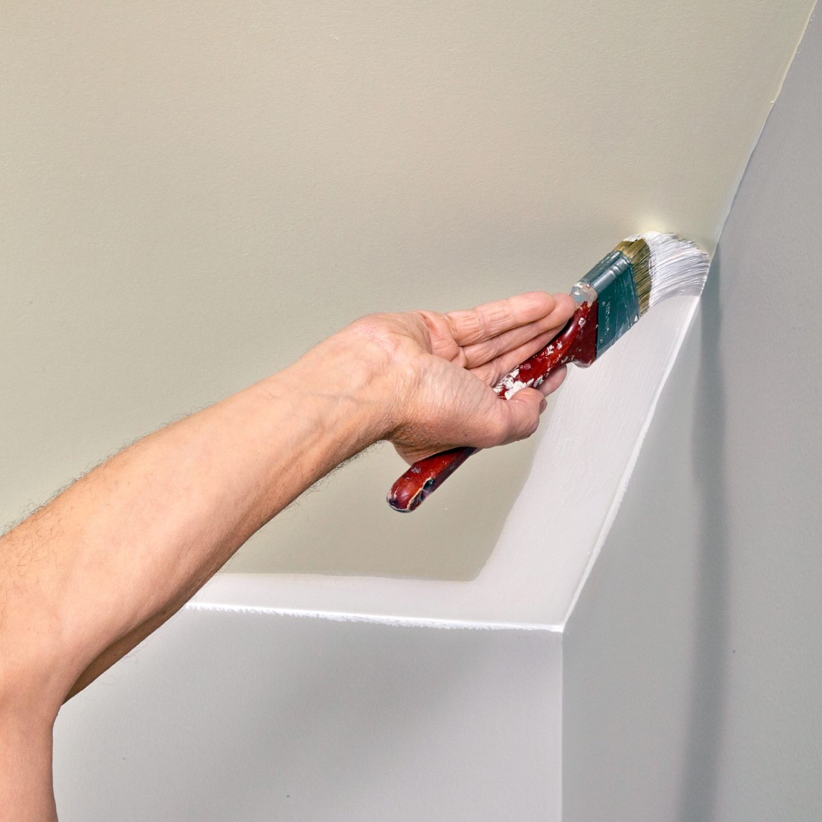 A person paints an interior wall corner with a brush. The wall is being painted white, and the person is holding a red-handled paintbrush, applying fresh paint along the corner edge. The arm and brush are the focus of the image.