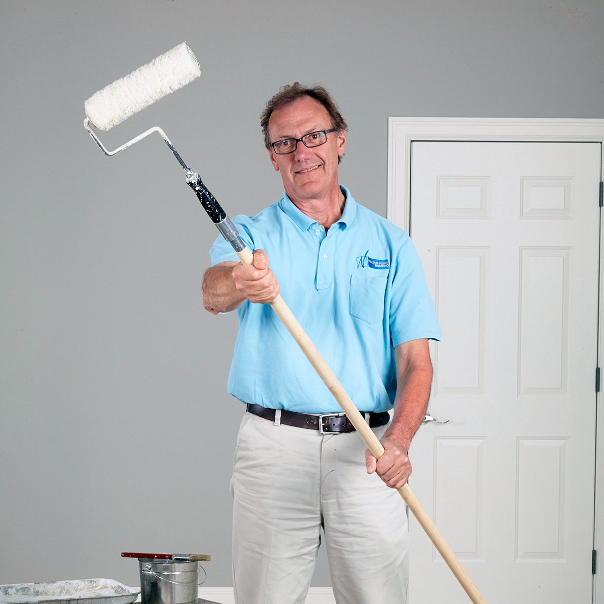 A person in a light blue polo shirt and white pants holds a paint roller extended towards the camera. They are standing in a room with a closed white door and a gray wall, with painting supplies on the floor.