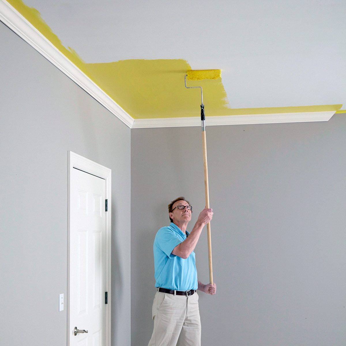 A person in a blue shirt and beige pants paints a ceiling yellow using a long roller. The room has light gray walls and a white door.