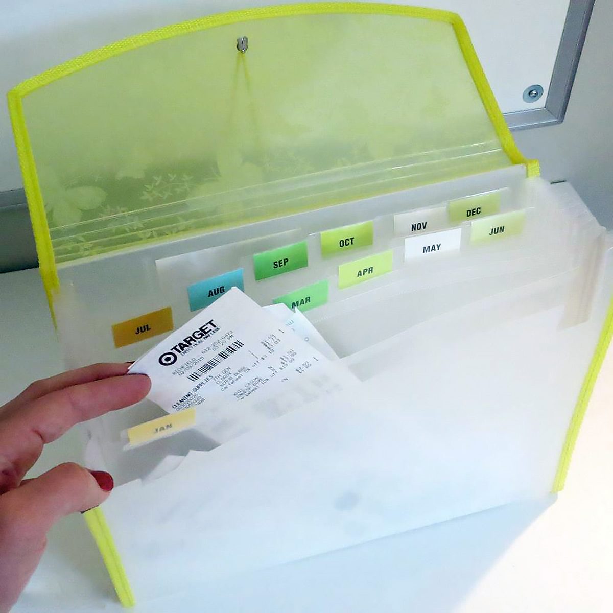 A person holds a receipt while organizing it into a translucent accordion folder with labeled dividers for each month. The outer edge of the folder is yellow, and the background is a white surface.