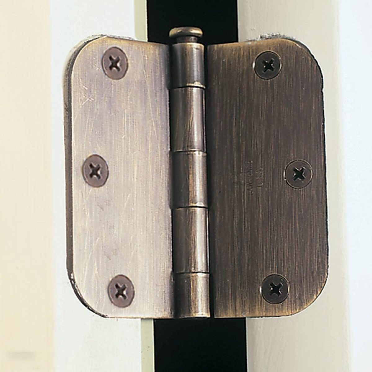 A close-up of a metal door hinge attached to the edge of a white door. The hinge is held in place by four screws, two on each side, with a brushed bronze finish. The door is slightly ajar, showing the hinge