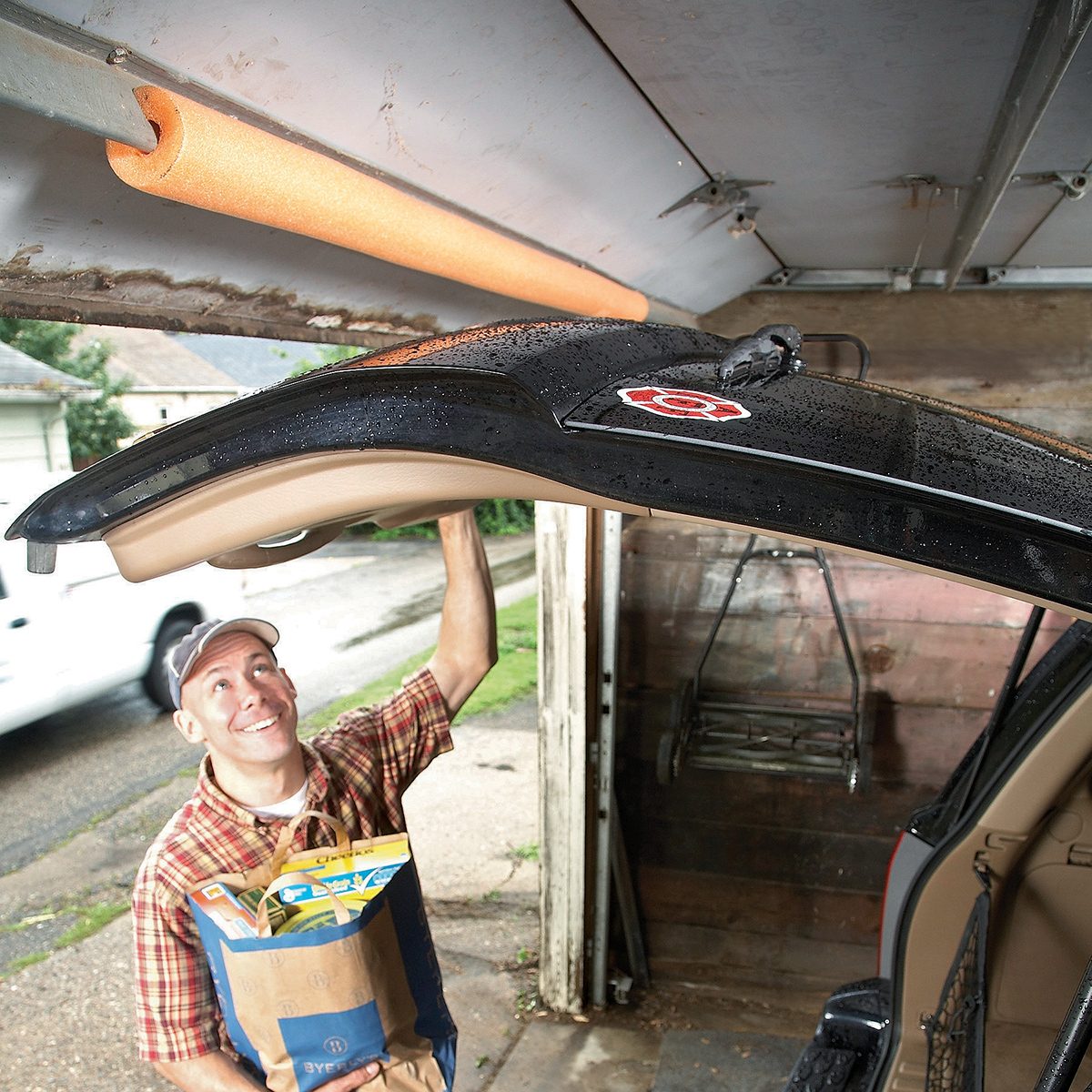 A person holds the open hatch of a car in a garage. They are smiling and wearing a cap and plaid shirt. The garage door has a foam noodle attached as a protective measure. They are holding several grocery bags.