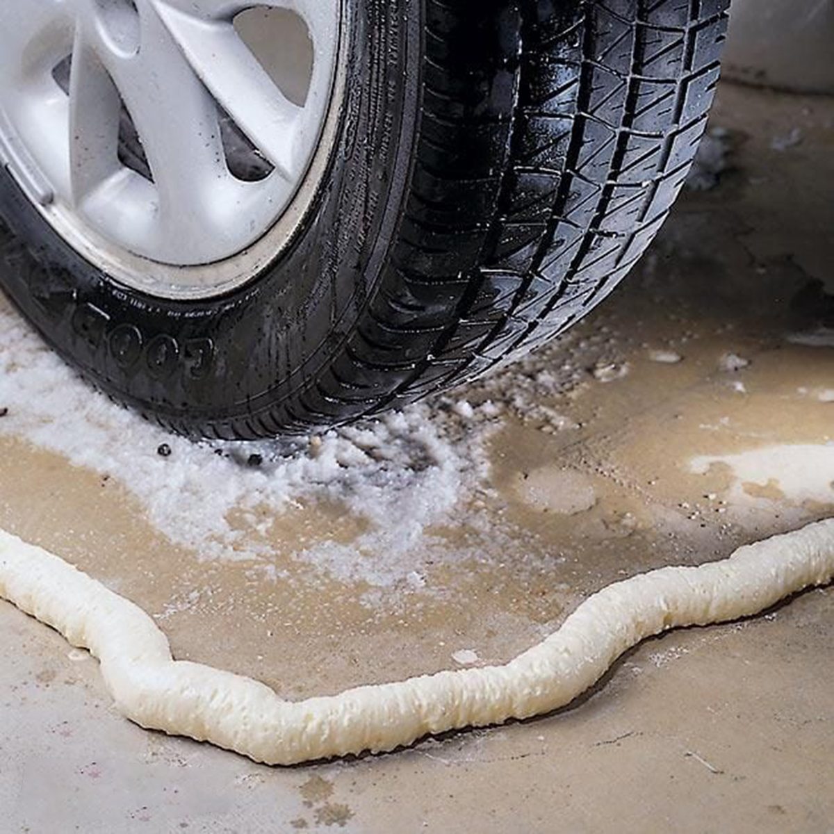 A car tire is parked in a garage with a strip of expanding foam sealing the floor. The foam appears to be preventing water from flooding into the space. The concrete floor shows signs of moisture and puddles.