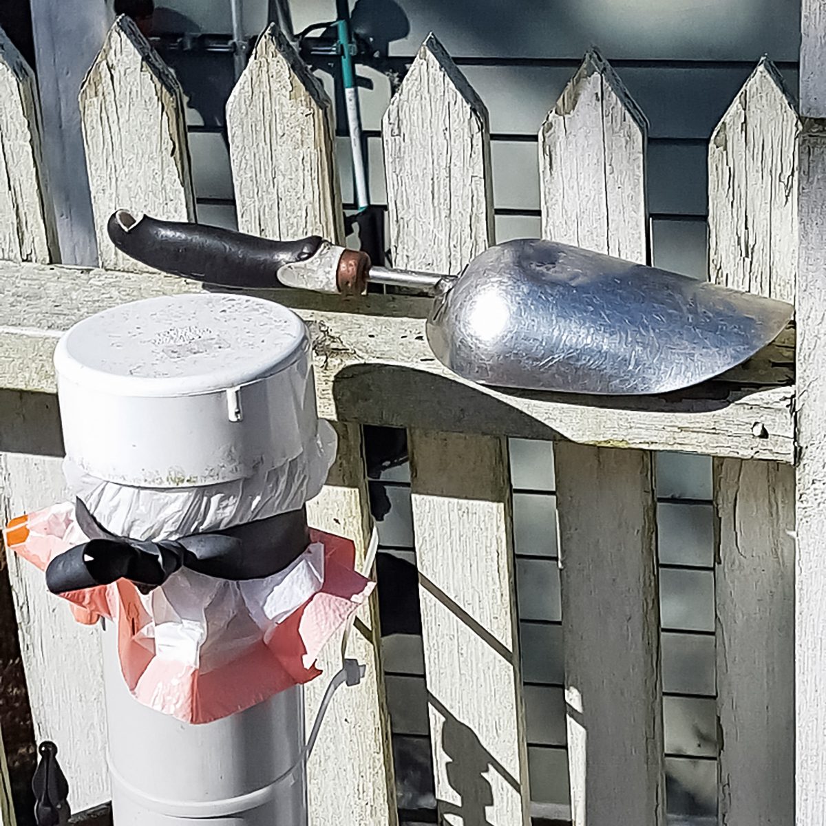 A metal gardening trowel rests on top of a weathered white picket fence. Next to it is a covered cylindrical object, possibly a pipe or vent, adorned with a black ribbon and a partially visible orange and white cover.