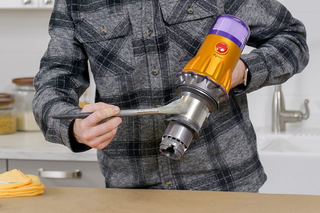 A person in a plaid shirt holding a vacuum cleaner and dipping a spoon into its nozzle over a kitchen counter. A sink and jars are visible in the background.