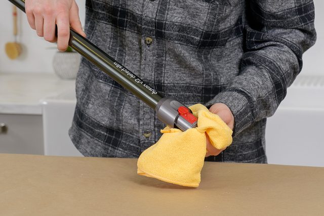 A person in a plaid shirt uses a yellow cloth to clean a vacuum cleaner handle. The scene is indoors, with a beige surface and a blurred white background.