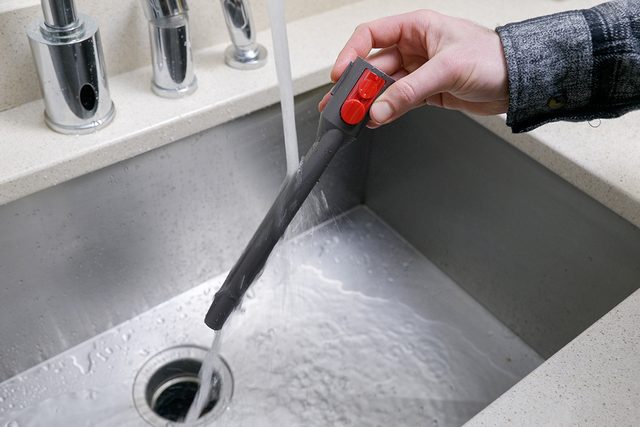 A person holding a grey faucet sprayer with red buttons, rinsing water into a stainless steel sink. The countertop is light-colored.